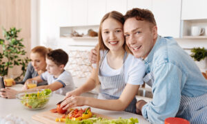 En familj med två vuxna och två barn står vid köksbänken och skär grönsaker